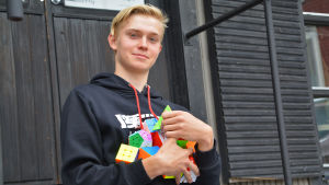 Famnen full av pusselkuber. Närbild på en tonårig pojke utanför ett svart trähus. Pojken håller en massa färggranna Rubiks kuber i sin famn och tittar in i kameran.