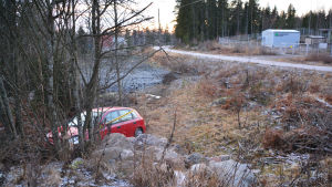 Bilen plöjde genom åkern en bra bit innan den stannade. En liten röd bil med framändan på en stenhög på en åker. Bilen är lite tillknycklad framtill och har ett gult plastband om sig. Bakom bilen syns vägen en bit bort.