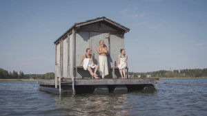Sauna Obscura kelluu Loviisanlahdella, saunomaan pääsee soutuveneellä Kolme pyyhkeisiin kääriytynyttä ihmistä seisoo harmaan kelluvan saunan edustalla ja katsoo merelle päin.