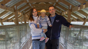 Maria och Mikael inne i ett av skugghusen. Här spenderar Mikael den mesta av sin tid. En man och en kvinna som håller att barn i famnen. De står mellan två långa rader med rävburar.