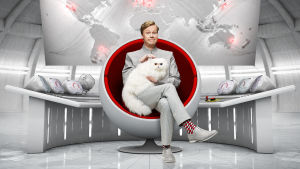 Johan Glans gästar ÅST den 12 april. Johan Glans sitter i en rund fotölj likt en skurk från en james bond film. han har en vit angorakatt i famnen.