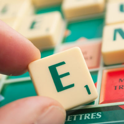 En hand håller upp bokstaven E på ett Scrabble-bräde.