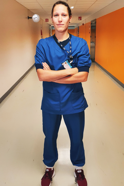 En intensivvårdare står i en sjukhuskorridor och tittar rakt in i kameran. Hon har blå jobbkläder på sig och armarna i kors.