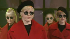 Nainen punaisessa takissa, pyöreissä aurinkolaseissa ja musta baskeri päässään.