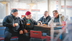 Neljä eläkeläismiestä istuu huoltoaseman pöydässä kahvilla ja keskustelee hymyillen.