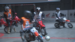 Neljä pelaajaa minicrosspyörien selässä tavoittelee jalkapalloa. Motoball-pelaajilla motocross-kypärät päässä. Ajoasuen päällä joukkueiden kevytliivit: valkoiset ja oranssit.