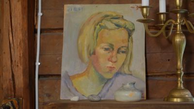 Hirseiseinä vasten nojaava valokuva nuorta Kirsi Kunnasta esittävästä maalauksesta, jonka vieressä on kynttelikkö.