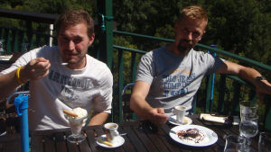 Kaksi viiksekästä miestä ravintolan terassilla nauttimassa jälkiruokia ja kahvia.