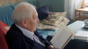 Lars Huldén sitter i en fåtölj och läser ur sina diktsamlingar