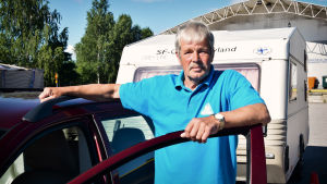 Olof Bussman ordnar Caramba-tävlingar i Finland. Olof Bussman i klädd en turkos pikéskjorta står lutad mot en bildörr. I bakgrunden skymtar en husvagn.