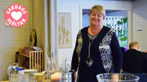 En bild på en kvinna i en sal, framför henne står mat på ett bord. I hörnet av bilden finns en stämpel där det står vardagshjältar. Kvinnan heter Mona Andersson.