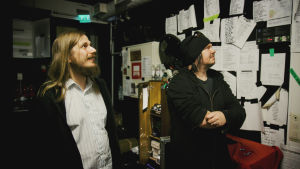 Kaksi miestä, Ville Karttunen ja Aki Tykki, katsovat ahtaassa tilassa seinille ripustettuja A4-kokoisia lappuja joihin on kirjoitettu eri bändien lauluja esitysjärjestykseen.