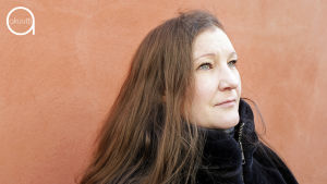 Riikka Sallinen seisoo oranssia seinää vasten.