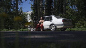 Med bilen kan Aapeli åka och klättra när han vill. En man sitter i förarsätet av en vit bil. Bilen är gammal. Förardörren är öppen.