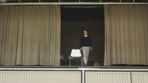 Henrik spelade hellre teater än fotboll. Han trivs fortfarande på scenen. En man går på en scen. Draperierna är halft fördragna och på scenen står en vit stol. Mannen går mot stolen.
