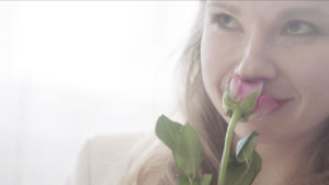 Anna-Riikka Rajanen roolihahmossaan haistelee vaaleanpunaista ruusua vaaleassa kuvassa.