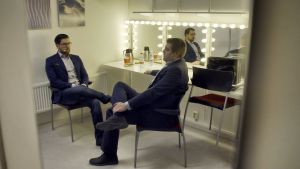 Jimmie Åkesson. Jimmie Åkesson sitter i en loge och väntar tillsammans med en annan man. Båda är klädda i mörk kostym och ser fundersamma ut. De sitter med ena benet upp på det andra.