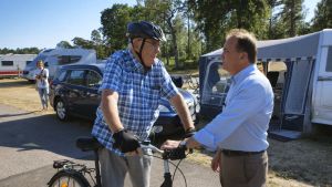 Stefan Löfven (till höger). Stefan Löfven är klädd i ljusblå skjorta. Han står på en kampingplats i sommarsolen och diskuterar med en äldre man som leder en cykel