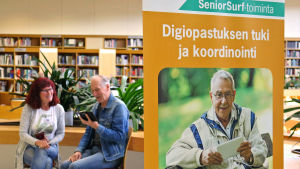Nettiä ikä kaikki -tapahtuman kaksi kävijää Kuopion kirjastossa, etualalla SeniorSurf-toiminnan juliste.