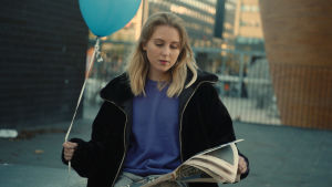 Molly vill visa upp både bra och dåliga dagar. En kvinna med långt blont hår, blå tröja och svart jacka. Sitter utomhus och på en stol och läser tidningen. Hon håller också i en blå heliumballong.