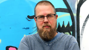 Kuvassa on kirjailija Tuomas Kyrö purkutuomion saaneen Kaurialan yläasteen, graffitin peittämän, seinän edessä.  