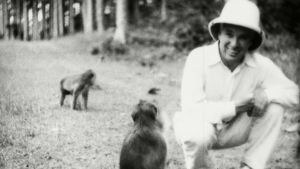 Chaplin Balilla. Charlie Chaplin kyykyssä hiekkatiellä, jossa kaksi apinaa. Katsoo kameraan.