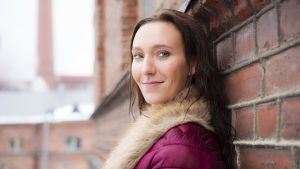 Saija Viherlaakso nojaa ulkona tiiliseinään, hymyilee.