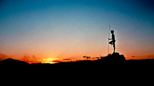 Nuori mies lannevaatteessa seisoo profiilissa aavikolla vasten auringonlaskua. Kuva elokuvasta Walkabout.