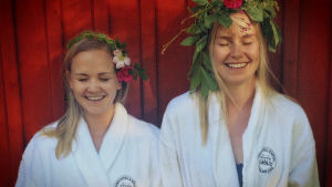 Kaksi vaaleaa naista nauraa silmät ummessa punaisen puutalon edessä kukkaseppeleen hiuksillaan.