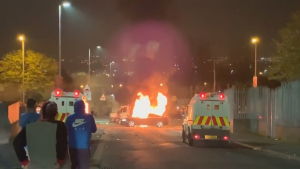 Både polisfordon och andra bilar stacks i brand under nattens kravaller i Londonderry. På bilden brinner en bil i londonderry. Tre åskådare syns i förgrunden.