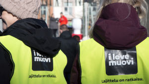 Kaksi henkilöä seisovat selin kameraan. Heillä on yllään keltaiset huomioliivit, joissa on I love muovi -kampanjapainatus.