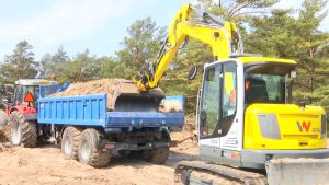 Traktor och släp med sand blir tungt på en sandstrand så det behövs lite hjälp för att ta sig fram. En grävmaskin puffar på en traktor med sand på släpet.
