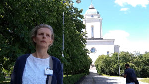 - Enligt lagen kunde du bli avrättad om du hade oäkta barn på 1700-talet. Men den lagen följdes sällan till punkt och pricka, berättar guiden Raija Lehtovaara. En allvarsam kvinna stor i framgrunden, i skuggan av några träd. I bakgrunden syns Sveaborgs kyrka.