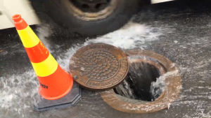 En massa vatten flödar in i en gatubrunn. Brevid den står en trafikkon. En buss kör förbi brunnen.