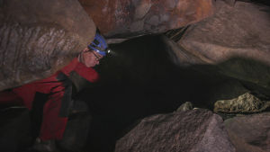 Punaiseen haalariin, siniseen kypärään ja otsalamppuun pukeutunut mies kävelee kumarassa kallioisessa tilassa.