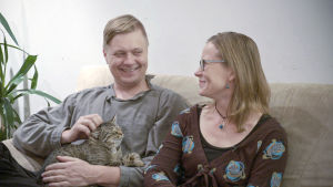 Ville ja Tiina Naumanen istuvat sohvalla. Villellä on sylissään kissa.
