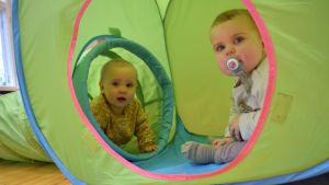 Tittut! Två babysar sitter i en grön leksakstunnel och kikar ut genom ingångshålet.