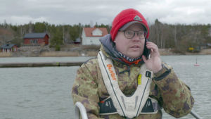 Jani Ollikainen beställer en sjöjungfrupizza. Jani Ollikainen, en man med röd mössa, grön jacka och flytväst, talar i sin mobiltelefon.