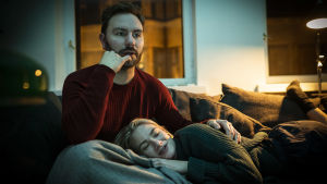 Mies ja nainen katsovat televisiota. Nainen makaa miehen sylissä, mies näyttää keskittyneeltä.