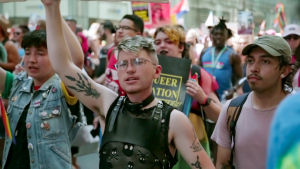 Nuori mies kävelee Pride-kulkueessa käsi ylöspäin osoittaen, mahdollisesti pitäen kylttiä.