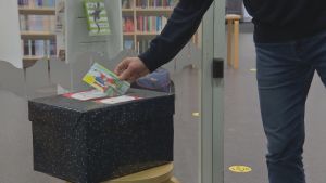 Man kan posta så många julkort som man vill till äldre. En hand sätter in ett julkort i en svart låda för att senare delas ut till ensamma äldre. I bakgrunden syns biblioteksböckr på hyllor.