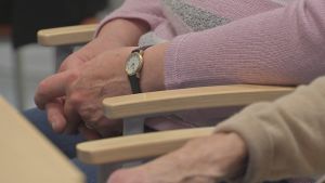 Många känner sig ensamma i dessa coronatider. En person med rosa tröja och armbandsur sitter med knäppta händer vid ett bord.