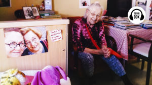 Aili istuu pöydän ääressä, vieressä olevan kaapin ovessa kuva Ailista Tuula-tyttären kanssa ja lappu jossa lukee "olen nyt Variston hoivakodissa".