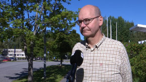 Heikki Kaukoranta säger att vi får vänja oss vid att han upp till hundra nya fall per dag. En flintskallig man med glasögon och rutig skjorta står i solen med en mikrofon framför sig.