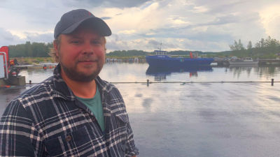 En man i keps står vid en hamn med en stor fiskebåt i bakgrunden.