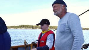 Tom Westerlund visar en skolelev från Tammerfors hur jakten ska styras. Tom Westerlund står vid rodret tillsmamans med en skolelev. Soligt försommarväder.