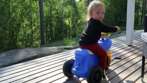 Blir det skola på svenska eller finska? Båda alternativen är möjliga även i lilla Dalsbruk. Ett litet barn på en blå tramptraktor, på en terass, skogen i bakgrunden.