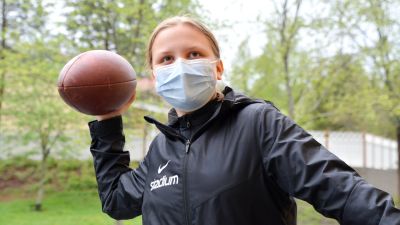 Julia Helppolainen i munskydd och svart jacka med en amerikansk fotboll i handen.