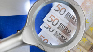 Bildsättning. Förstoringsglas över 50-eurossedlar, Finlands flagga i bakgrunden.