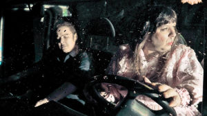 Pariskunta ajaa asuntoautoa sateessa. Kuvassa ohjaamo, jossa istuu mies ja nainen. Kuvassa näyttelijät Angelika Meusel ja Kai Tanner.